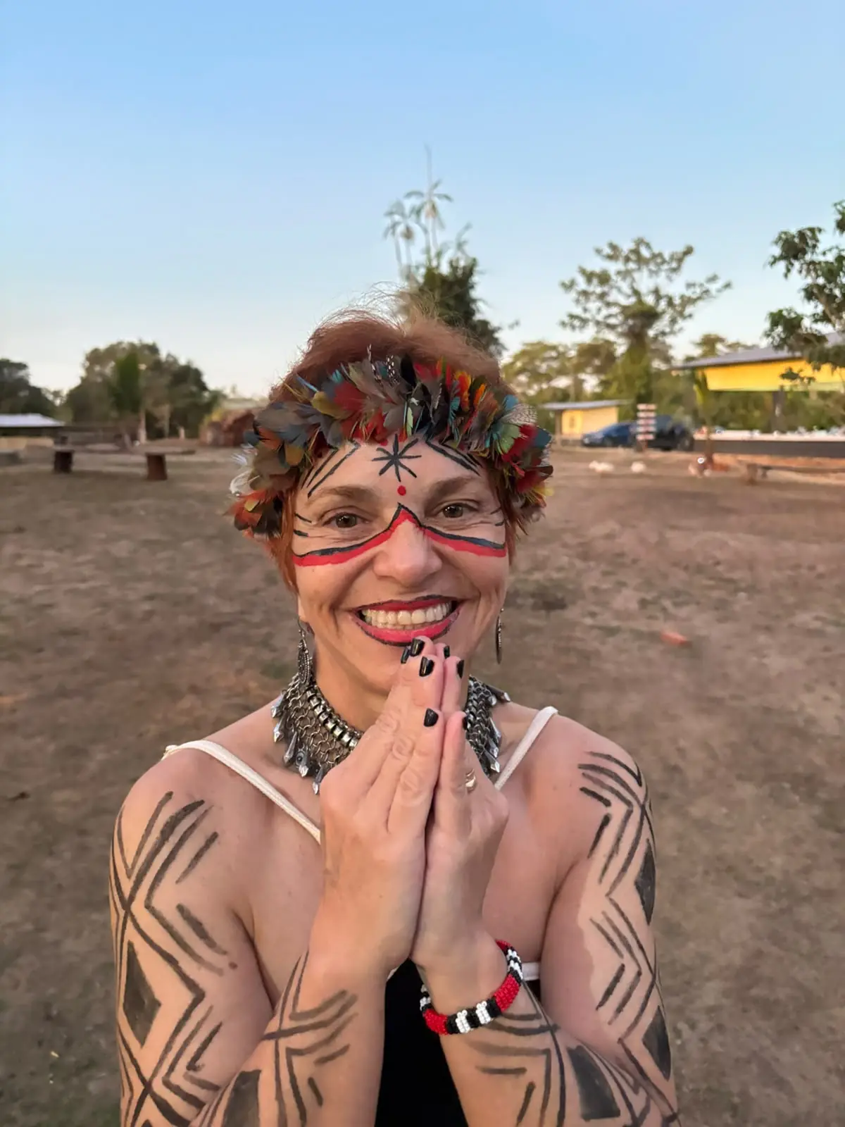 Sita Madhu com pintura ritual indígena e cocar de penas, sorrindo com as mãos unidas em saudação