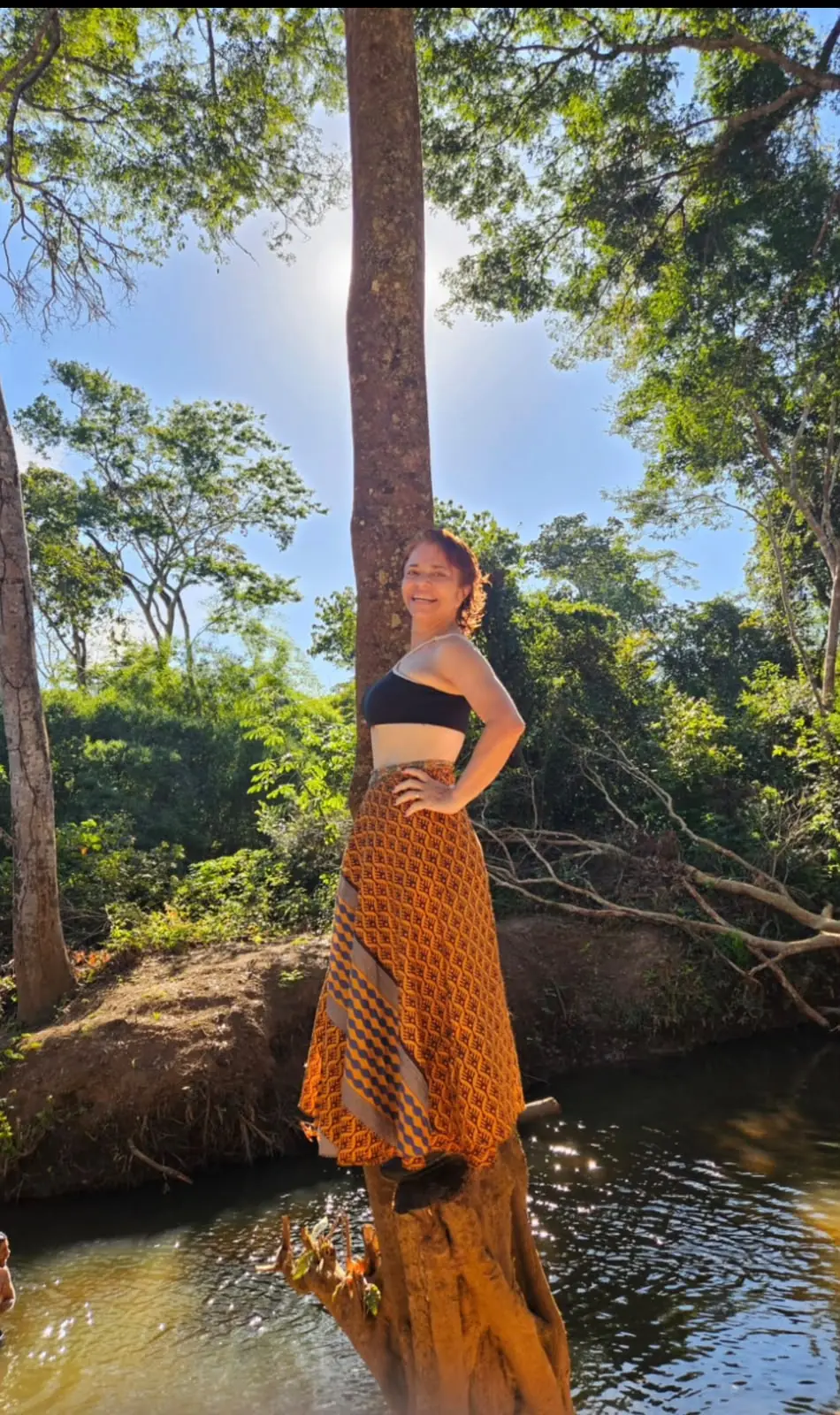 Sita Madhu sorrindo, com saia laranja, à beira do rio na Chácara Namastê