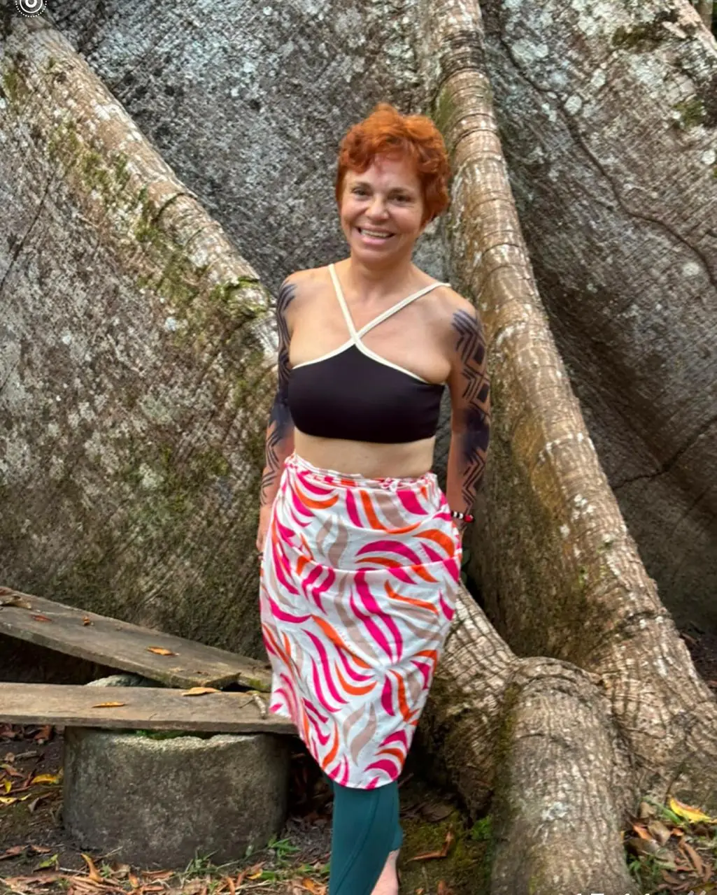 Sita Madhu sorrindo entre grandes raízes de árvore, vestida com saia estampada