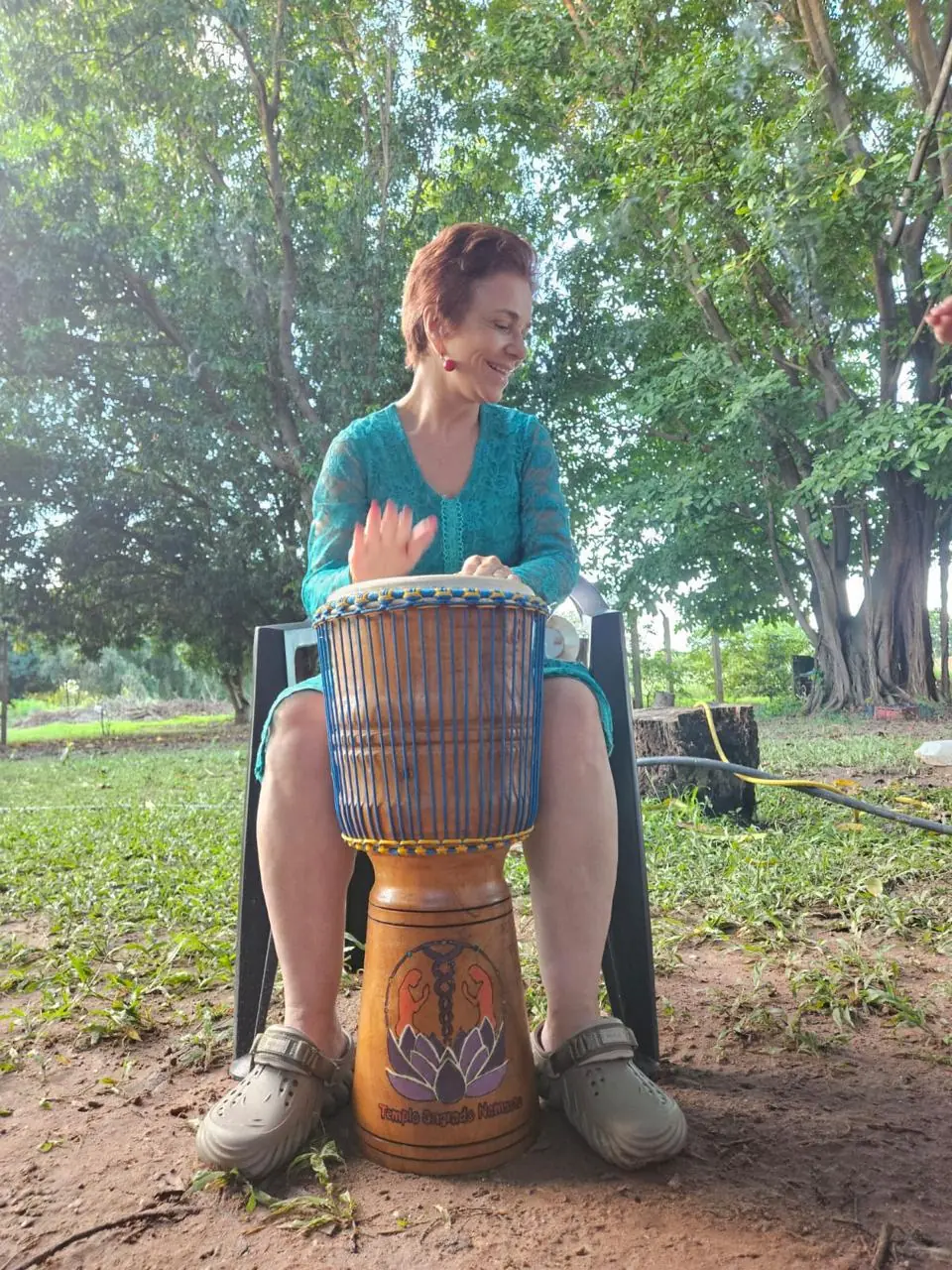 Sita Madhu tocando tambor, sorrindo, ao ar livre entre árvores